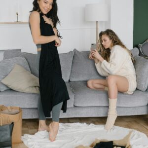 Two women relaxing while trying on new clothing purchases in a cozy living room setting.